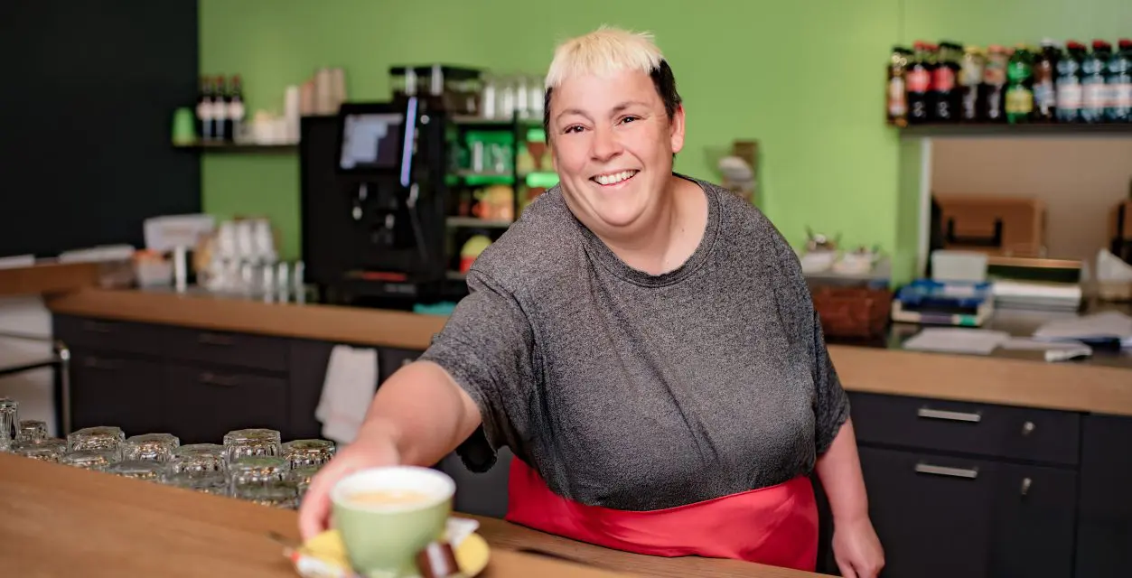 Frau hinter einer Theke die eine Tasse mit Kaffee entgegenstreckt
