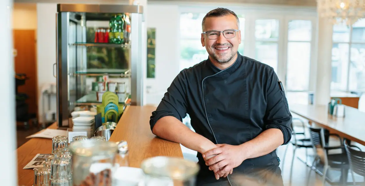 Porträt eines lächelnden Mannes. Er lehnt sich auf Theke im Hintergrund links eine Glasvitrine rechts im Bild ein langer Tisch mit Stühlen und im Hintergrund ein Fenster