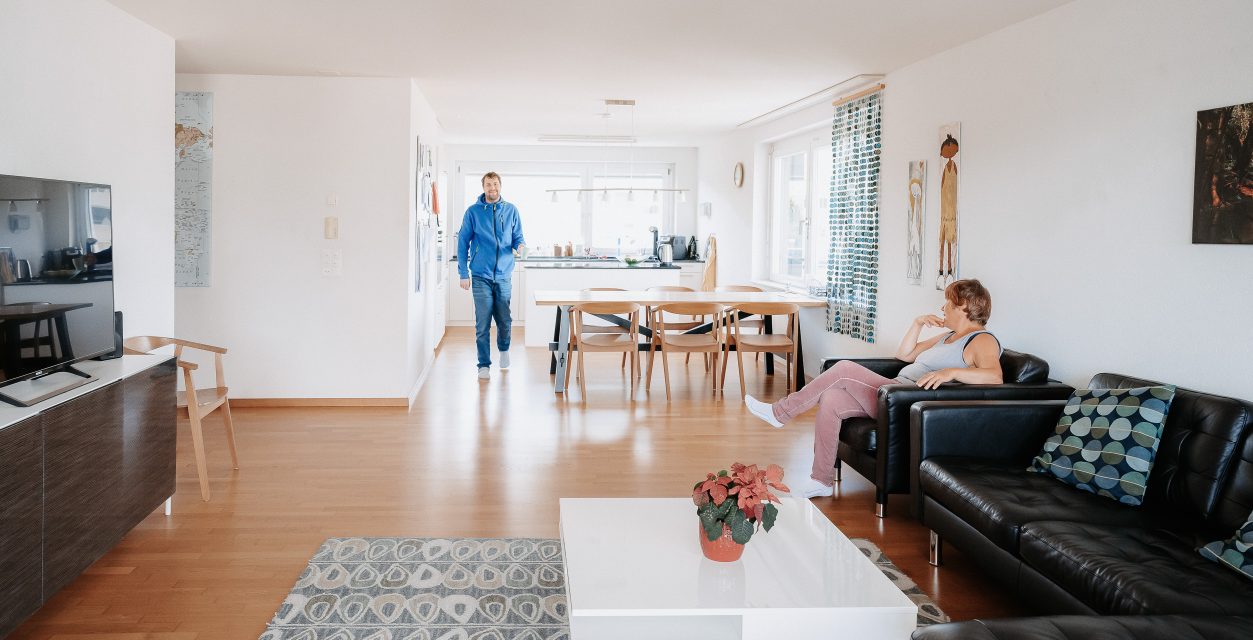 Ein Foto von einem Wohnzimmer Richtung offene Küche und Essbereich. Es sind sehr helle Räumlichkeiten. Ein Bewohner läuft gerade von der Küche Richtung Wohnzimmer und eine Bewohnende sitzt links im Bild auf einem schwarzen Ledersofa, dass an der Wand steht.