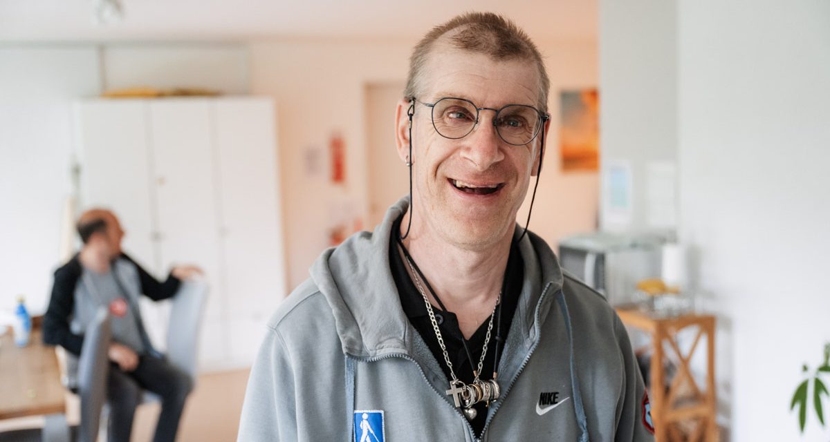 Porträt eines lächelnden Mannes mit Brille. Im Hintergrund verschwommen eine Esszimmertisch, an dem ein weiterer Mann sitzt.