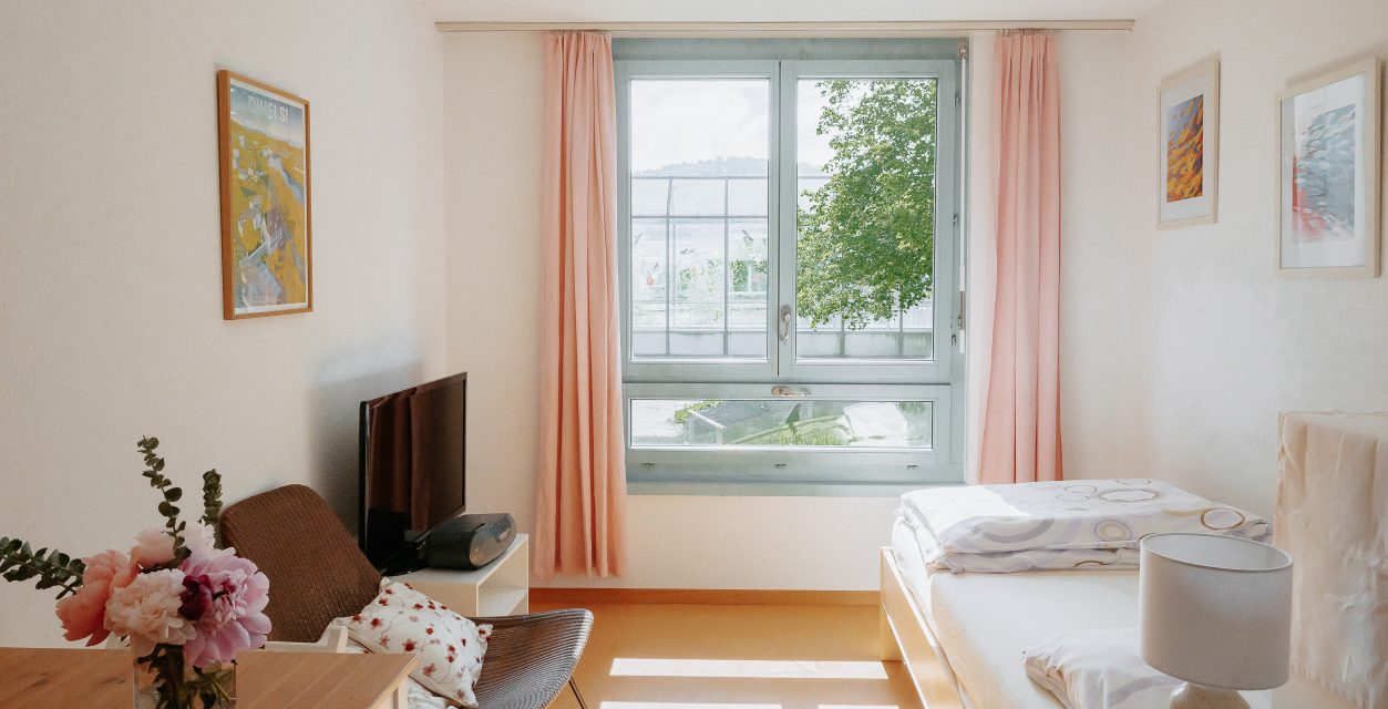 Ein helles Zimmer: in der Mitte des Fotos ist ein Fenster mit Blick ins Grüne mit rosafarbenen Vorhängen. Rechts steht ein Einzelbett an der Wand und links davon ein Stuhl und ein TV an der Wand.