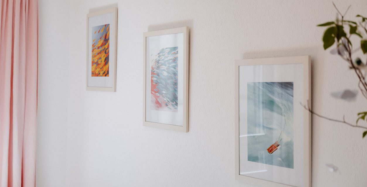 Eine Wand eines hellen Zimmers mit einem rosaroten Vorhang und drei gestuft aufgehängten Bildern.