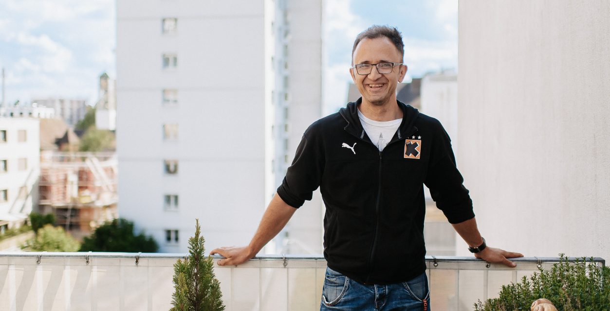 Porträt eines jungen, lächelnden Mannes mit dunkelen Haaren und Brille und schwarzem T-Shirt mit dem Kadetten-Logo. Er steht auf einem Balkon im Hintergrund Hochhäuser und grüne Umgebung.l