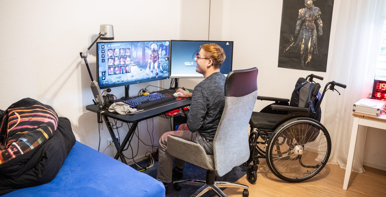 Porträt eines jungen Mannes im Bürostuhl vor einem Computer mit zwei Bildschirmen am "gamen". Rechts vom Tisch steht ein Rollstuhl