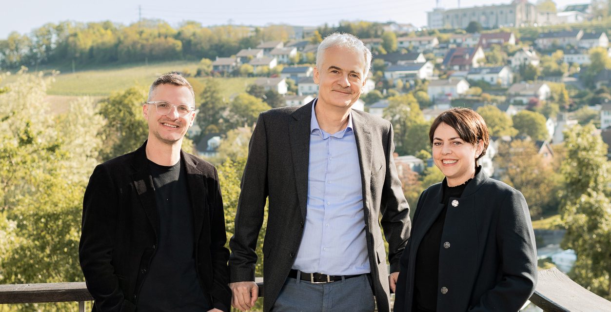 Porträt der Geschäftsleitung drei Personen von links nach rechts Thomas Ruhe-Zölle, Andreas Gutmann, Carmen Duss sie lehnen sich an das Geländer einer Terrasse im Hintergrund ist ein Fluss zu sehen sowie das ander Ufer mit Bäumen und Einfamilienhäusern.